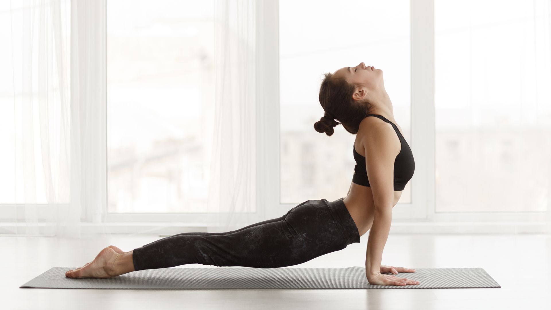 Woman in yoga pose, back arched, head up, in front of a window.