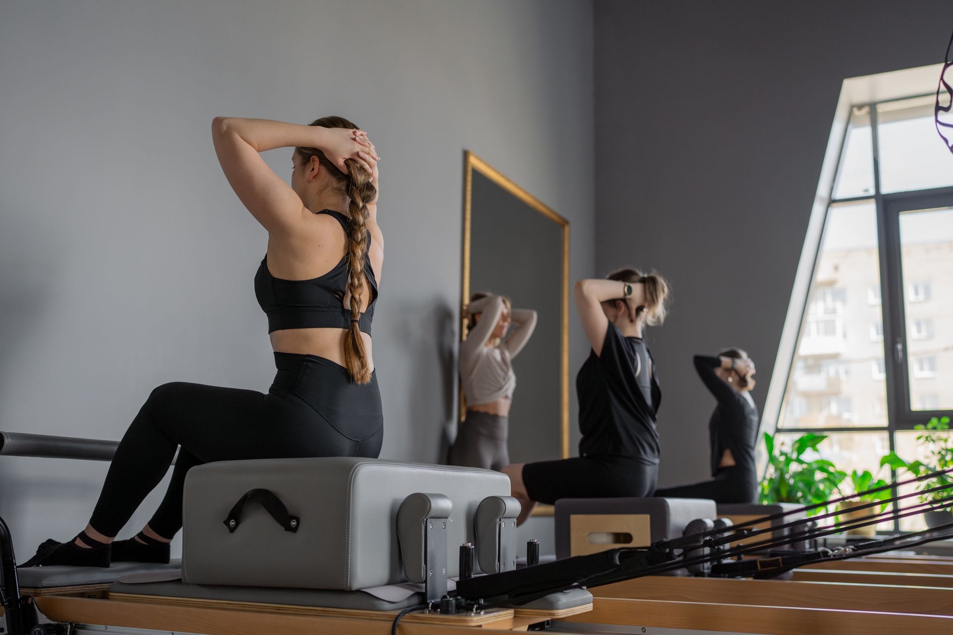 People doing pilates class, seated, hands behind heads, on exercise apparatus. Gray wall, window.