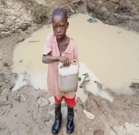 A young boy is standing in the mud holding a can of water.