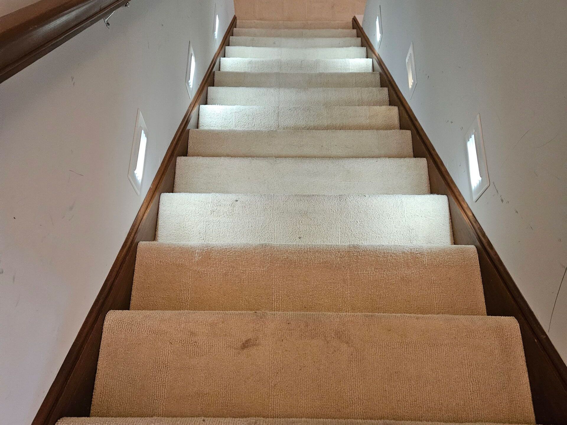 Staircase with beige carpet and wooden trim, lit by recessed lights on the walls.