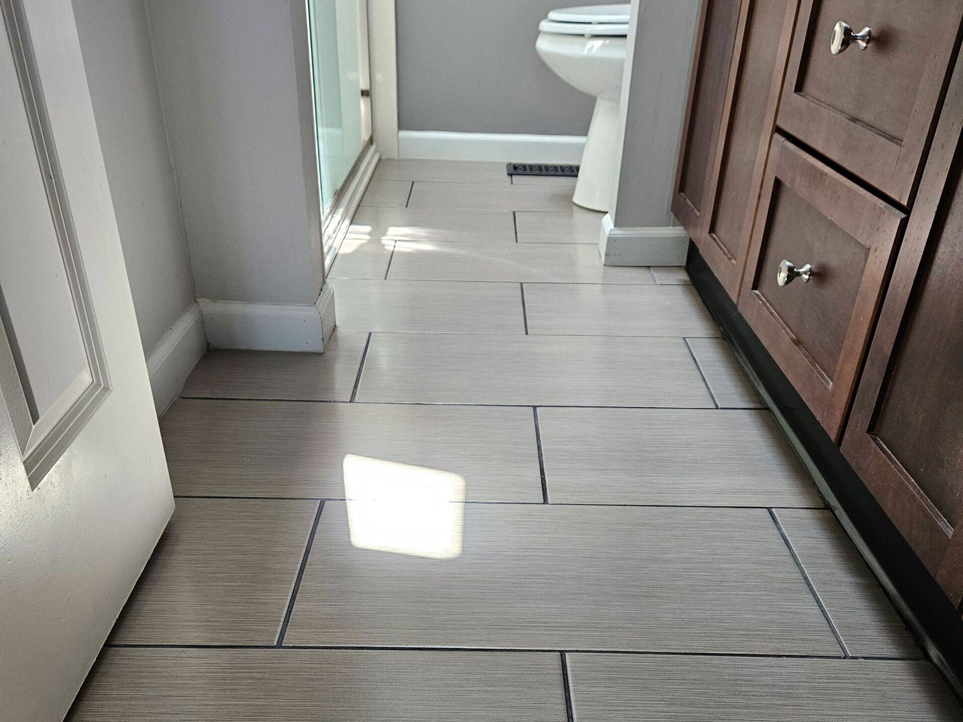 Bathroom with light-colored rectangular tiles on the floor, toilet, and wooden cabinets. Bright sunlight shines on the floor.