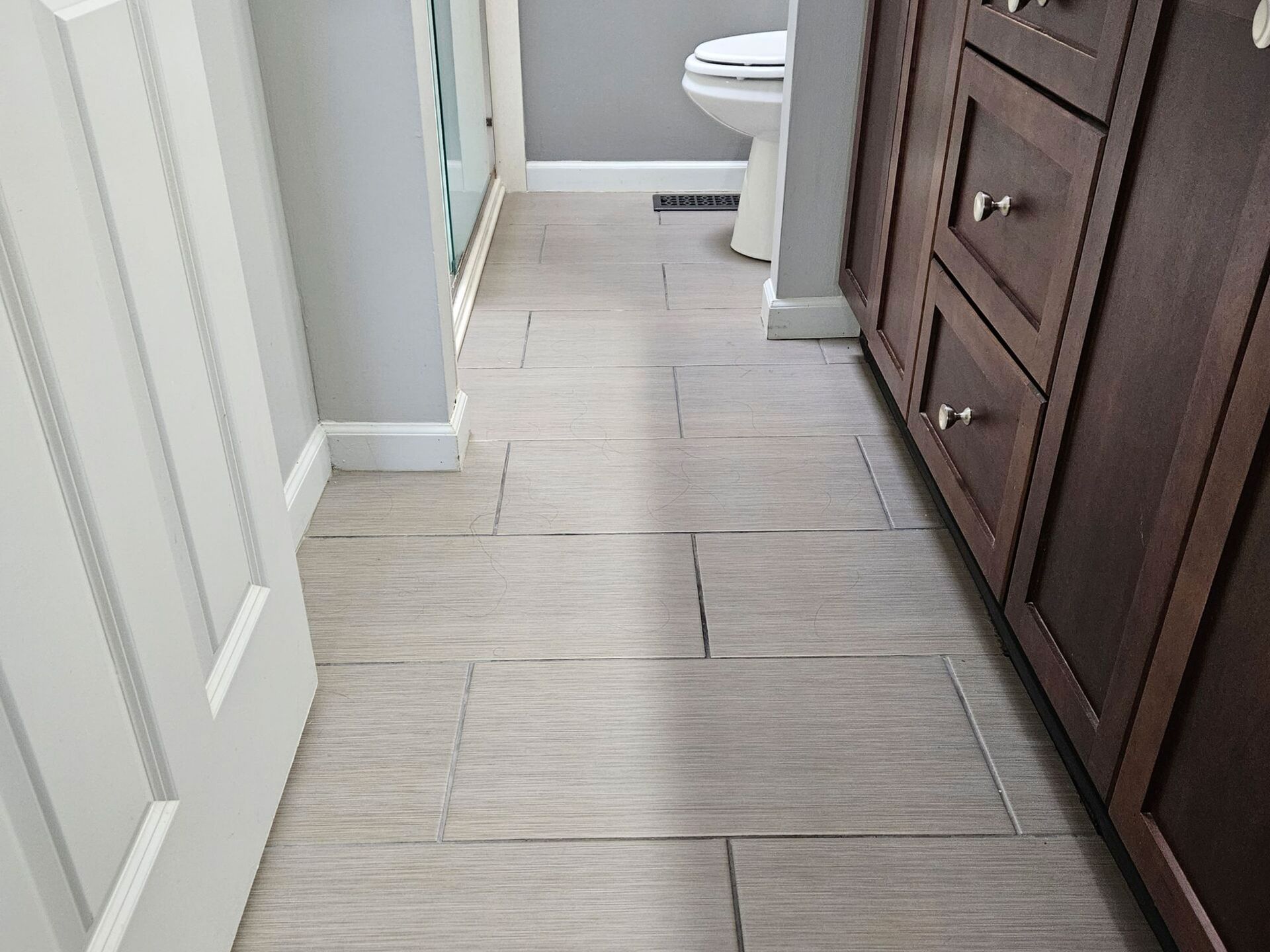 Bathroom floor with rectangular gray tiles, brown vanity on right, white door on left, toilet in background.