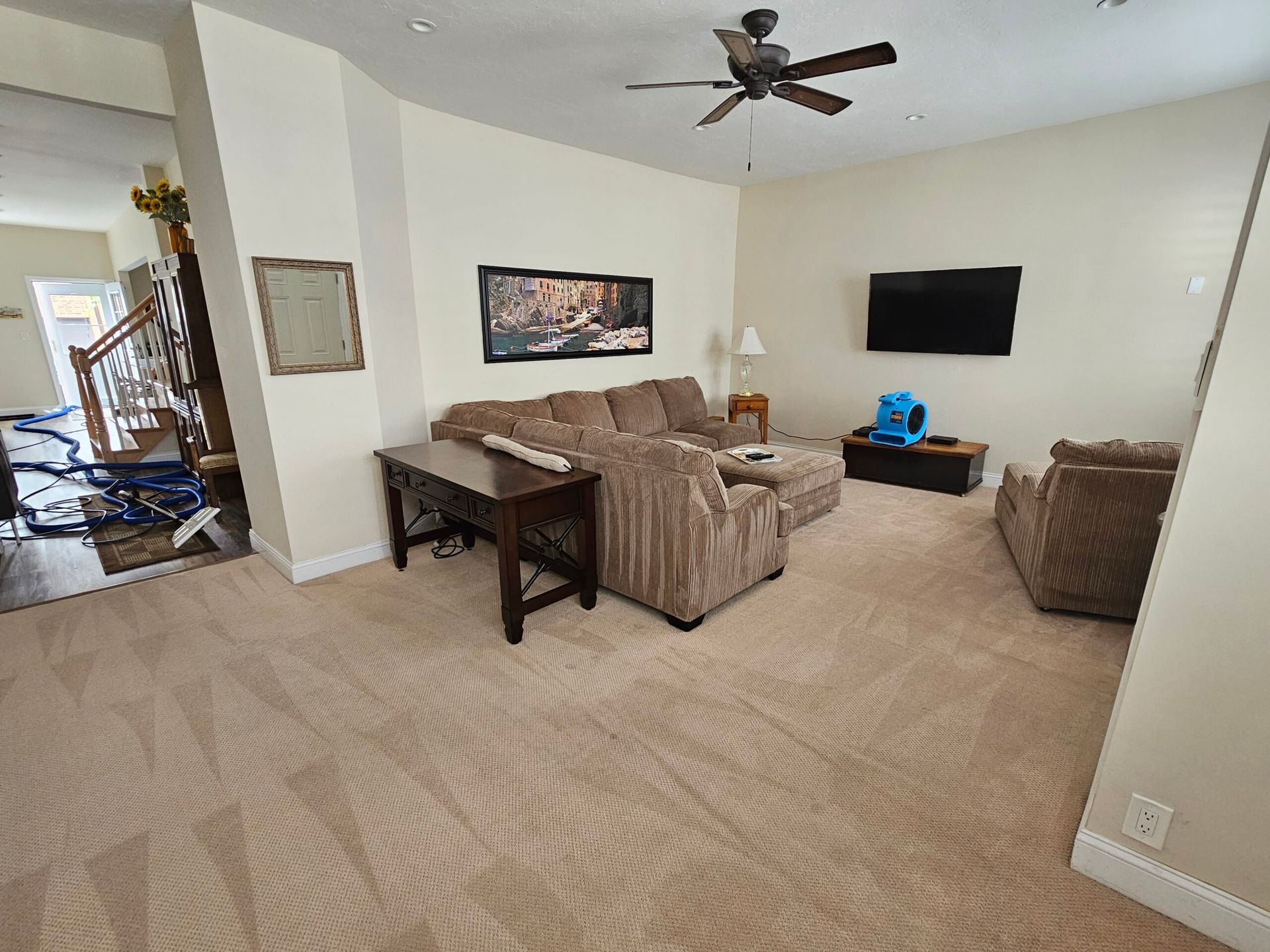 Living room with beige carpet, sectional sofa, TV, and a table; a fan hangs from the ceiling.