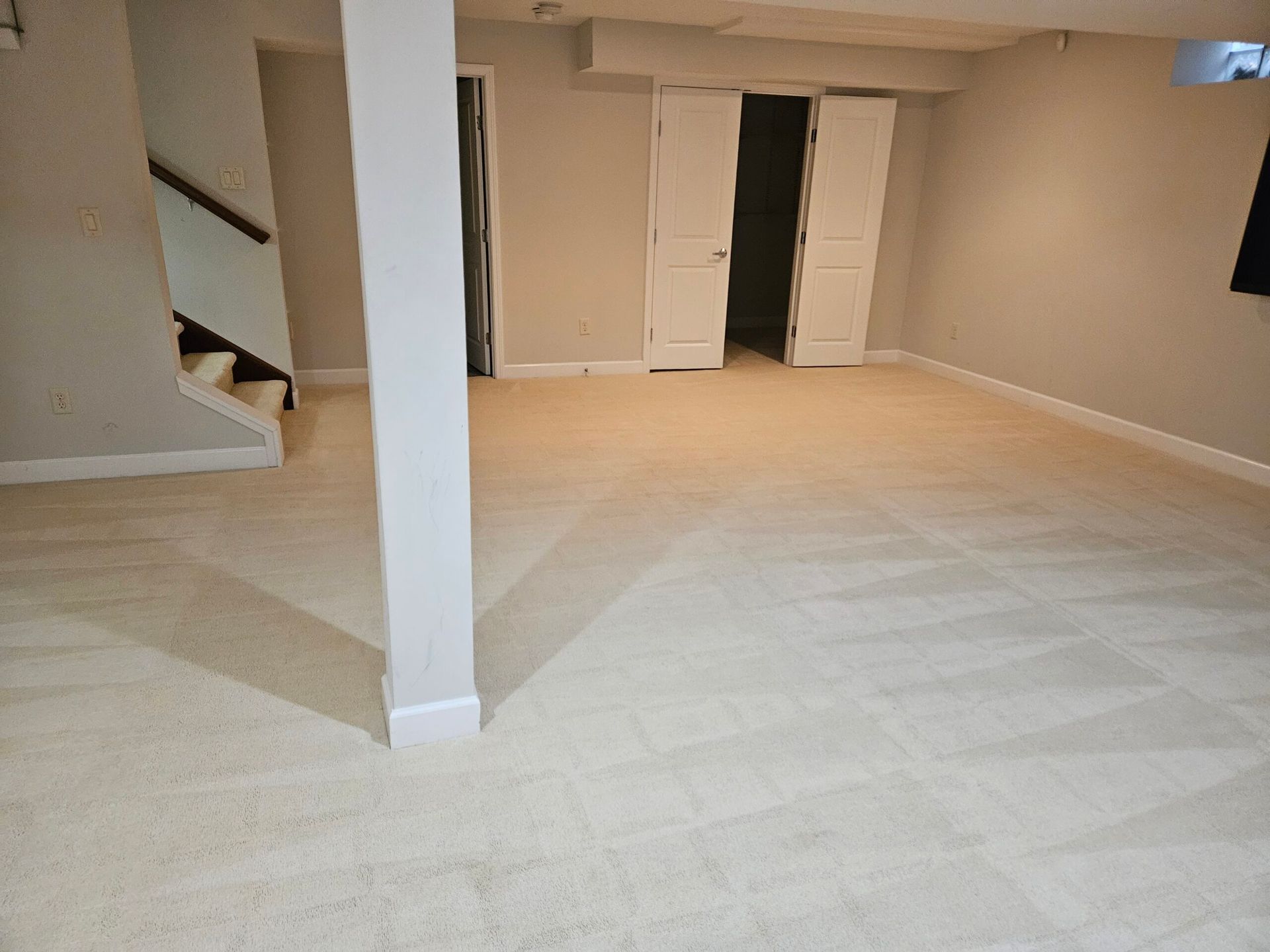 Empty basement room with tan carpet, white support beam, and staircase to left.