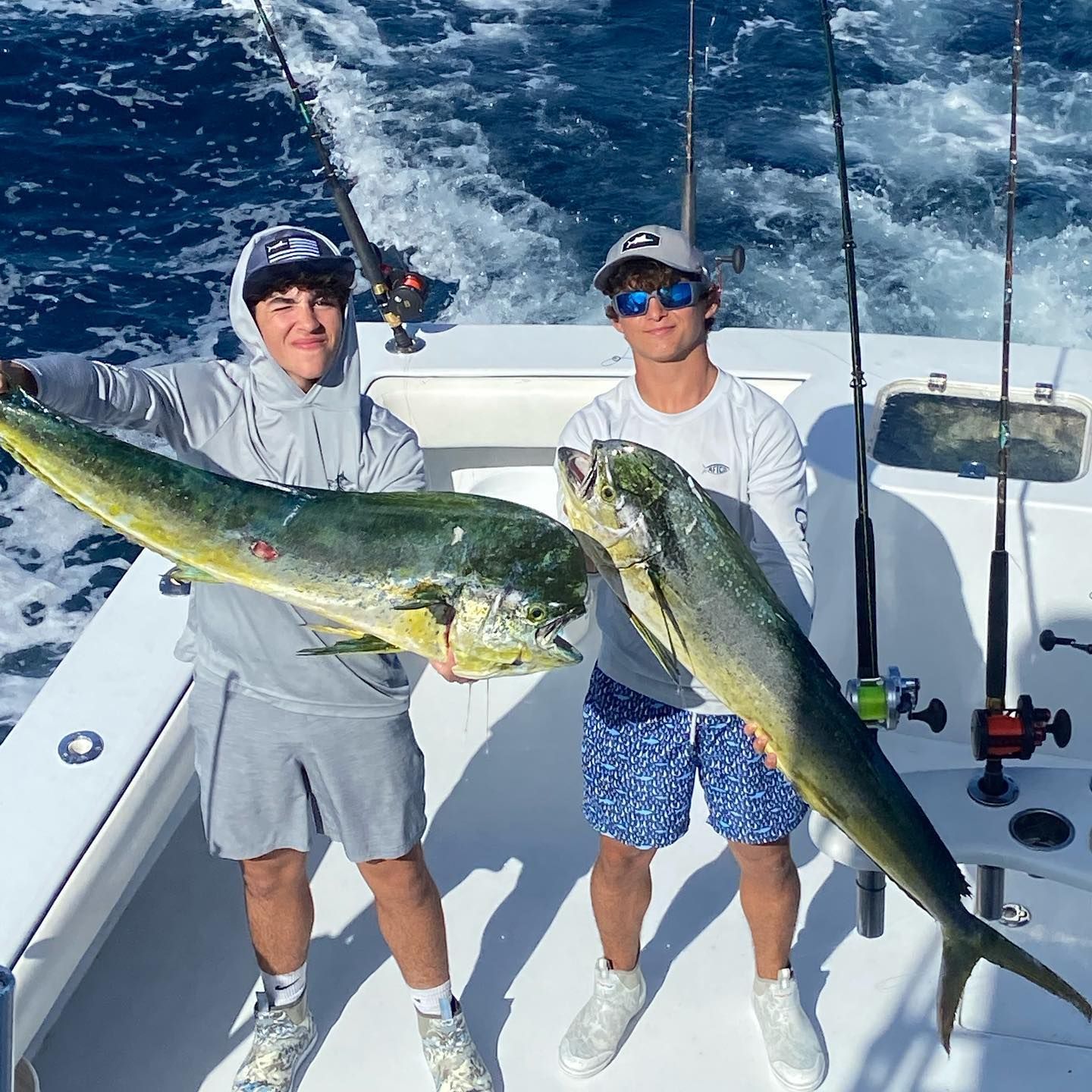 Two people on a boat holding large, colorful fish; ocean background, fishing rods visible.
