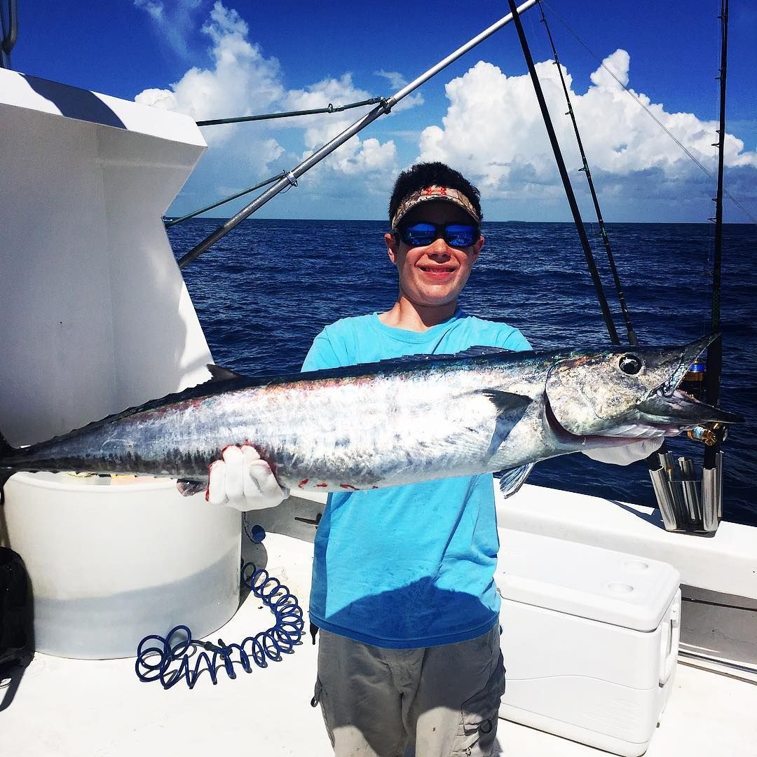 Young person on a boat holding a large fish they caught, blue water and sky.