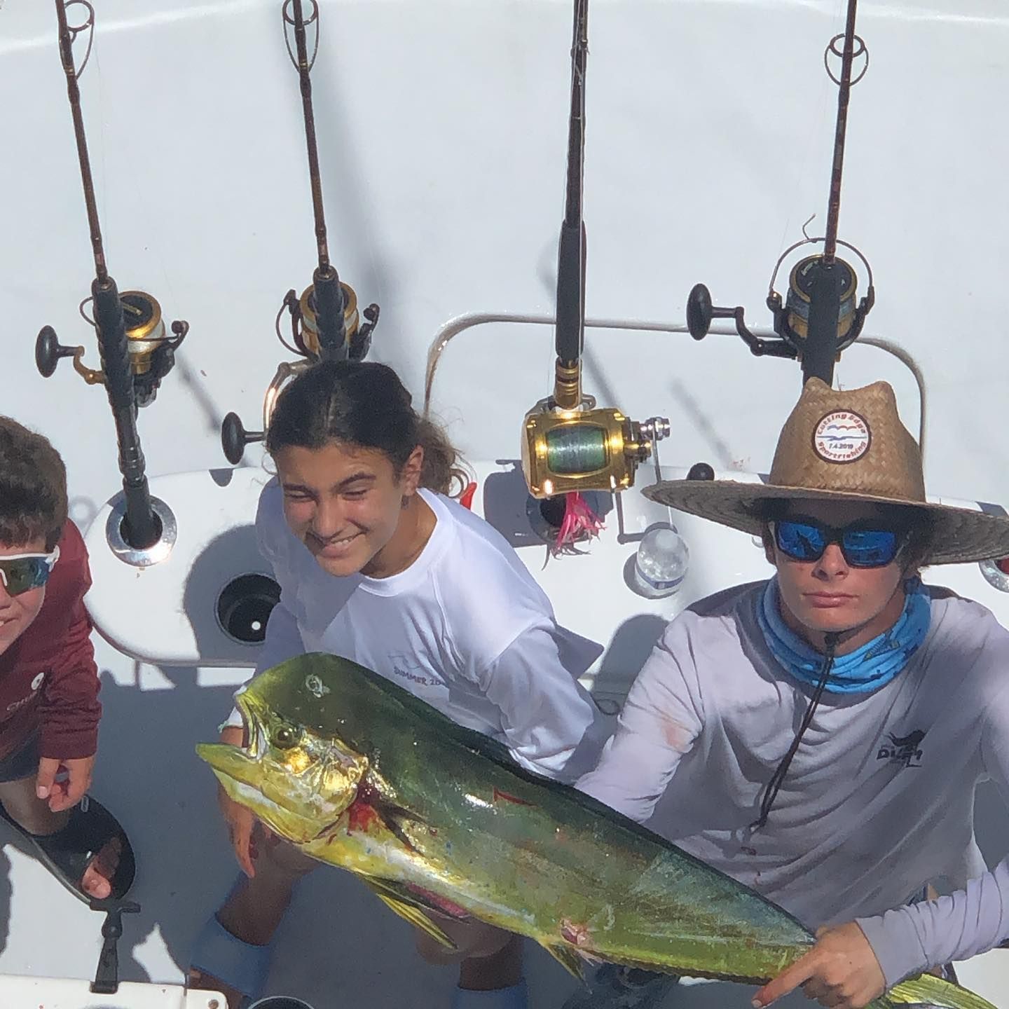 People on a boat with fishing rods holding up a large yellow and green fish; sunny, happy expressions.