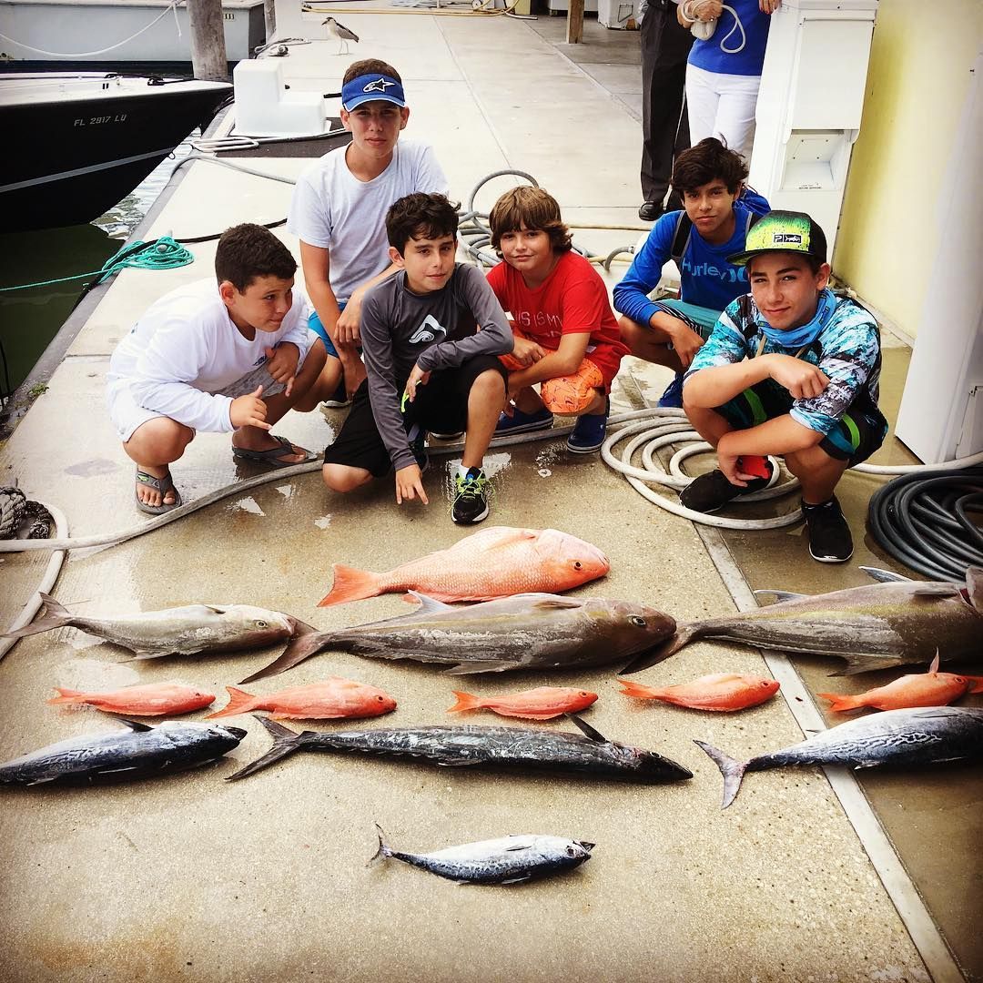 Boys crouch with fish on a dock, a boat in the background. Several different fish laid out.