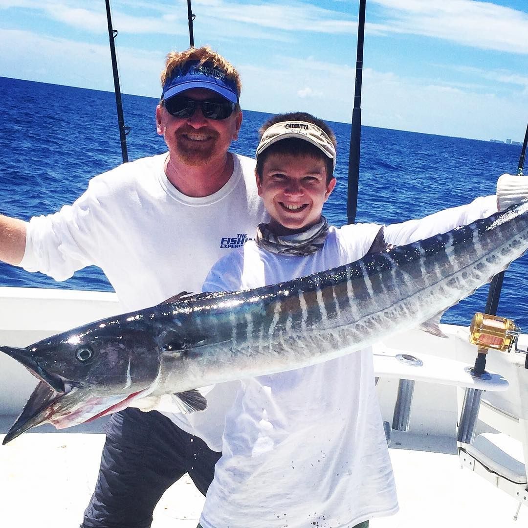 Man and boy smiling, holding large fish on a boat, blue water and sky.