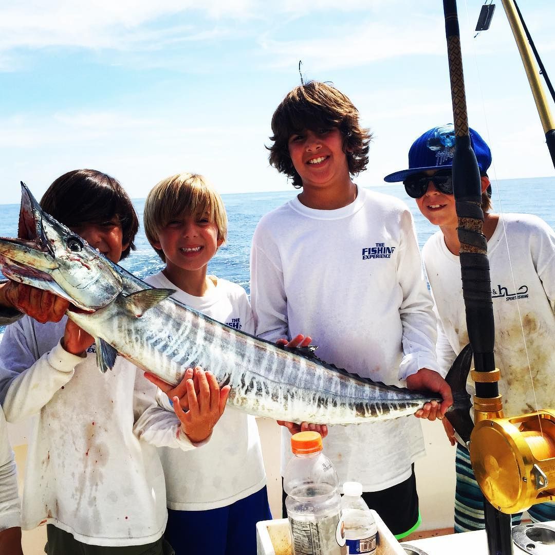 Four boys on a boat hold a large, silver fish, smiling. Ocean and fishing poles visible. Sunny day.