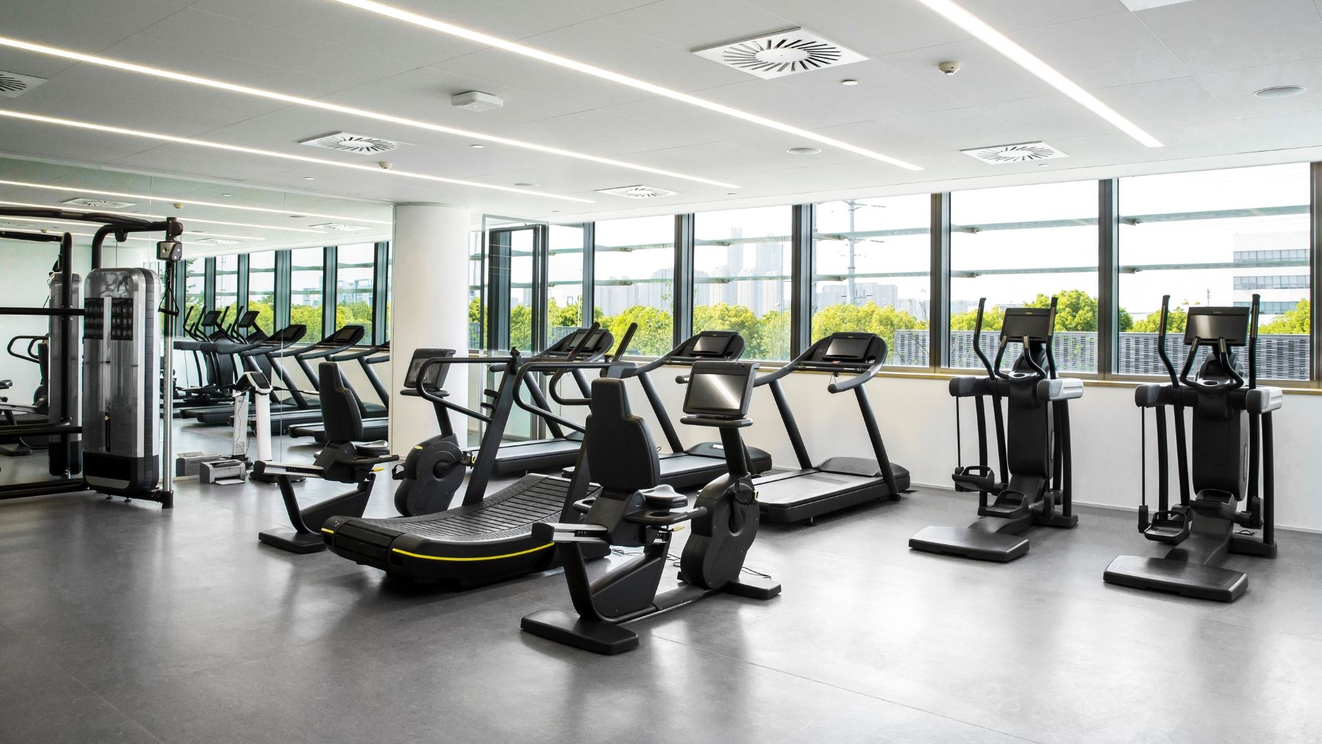 Gym with treadmills, elliptical machines, and weight equipment, with large windows overlooking a city.
