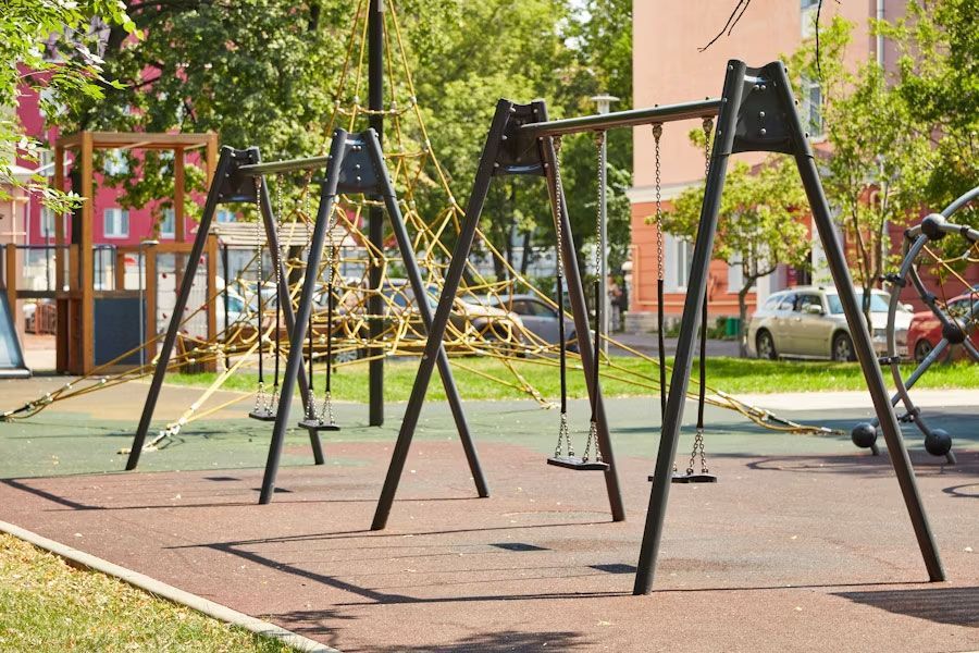 A playground with black swing sets on a green and brown surface, surrounded by trees and buildings.
