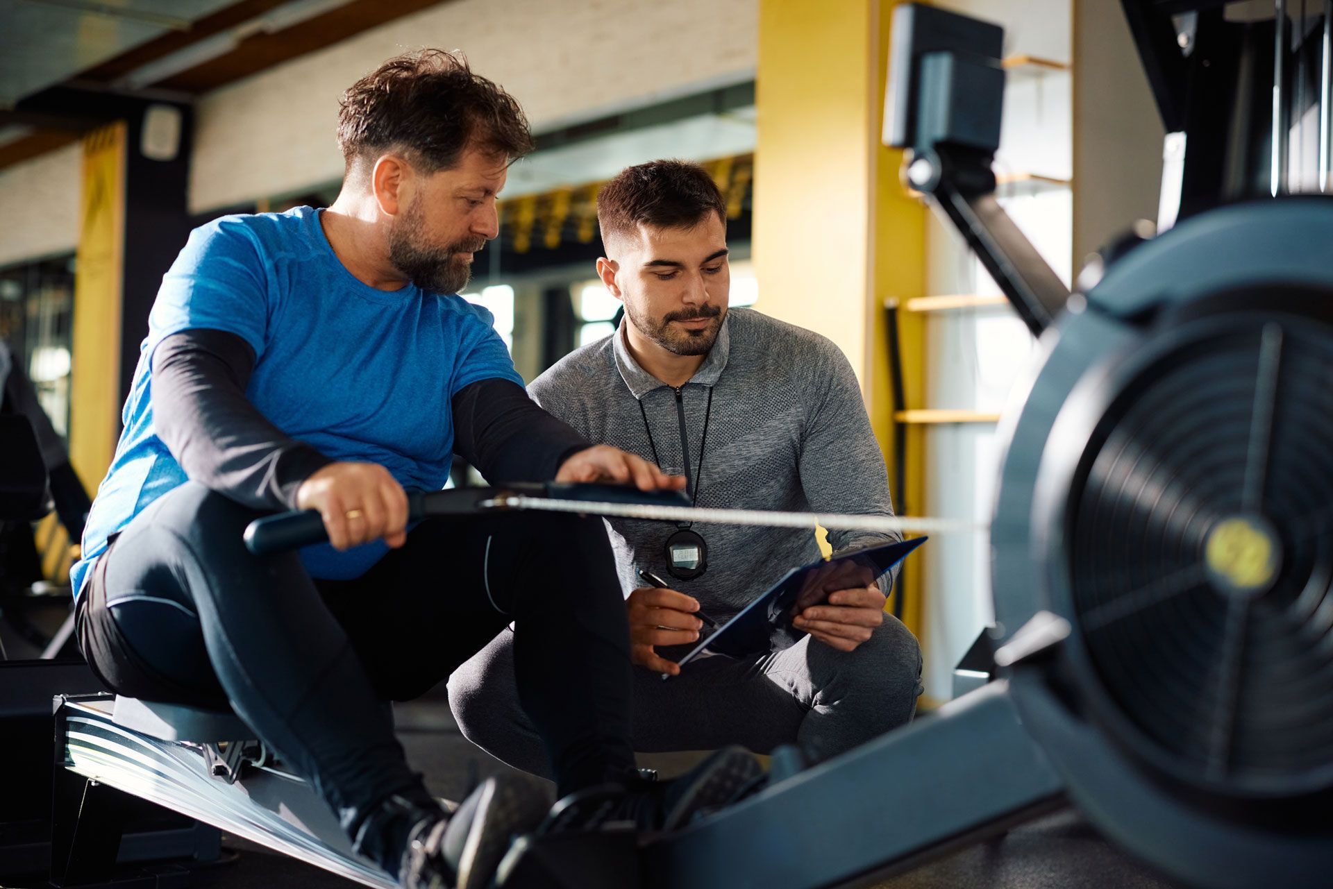 A man is riding a rowing machine in a gym while another man looks at a tablet.