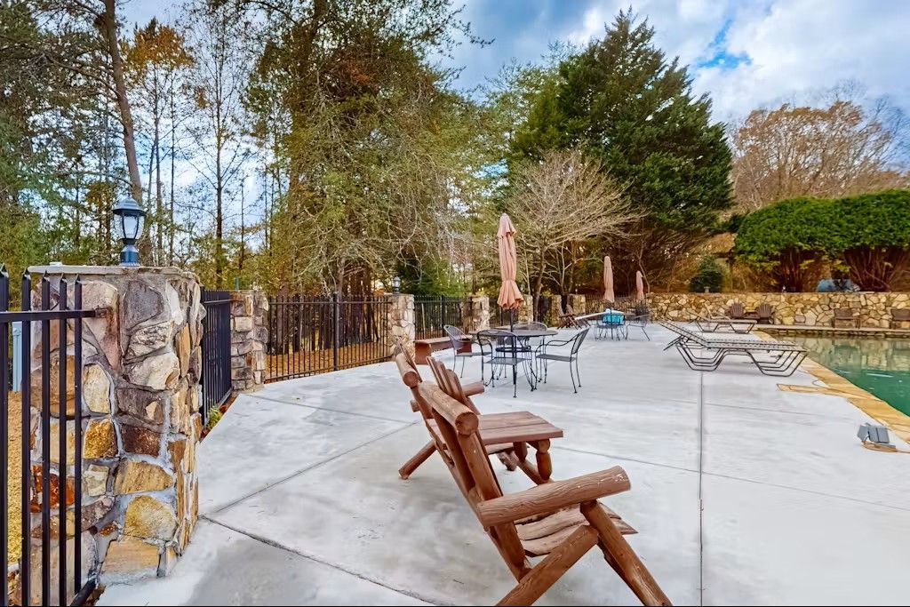 Patio with wooden chairs, tables, and a pool surrounded by trees under a cloudy sky.
