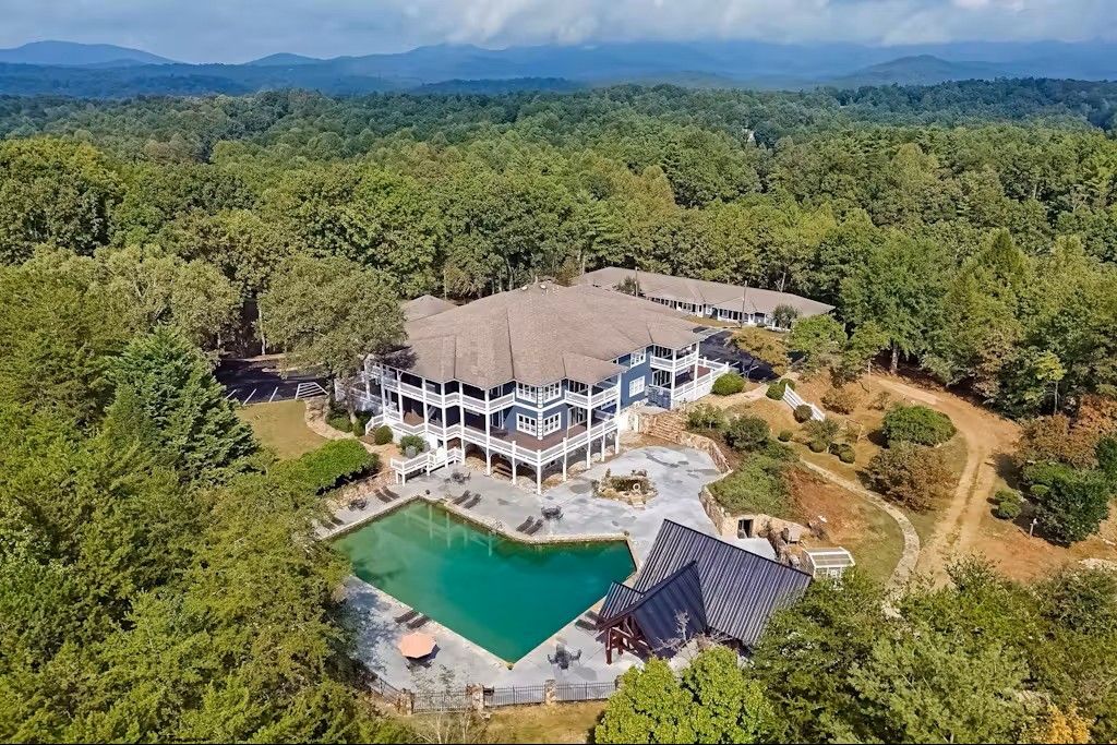 Aerial view of a large white house with a pool, surrounded by forest and mountains.
