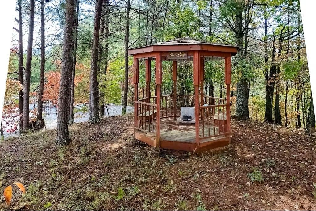 Wooden gazebo in a forest setting with a small structure inside.