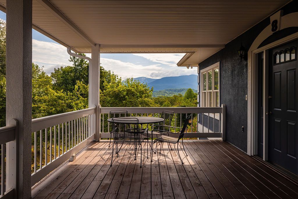 Covered porch overlooking trees and mountains; table and chairs, dark blue house, sunny.