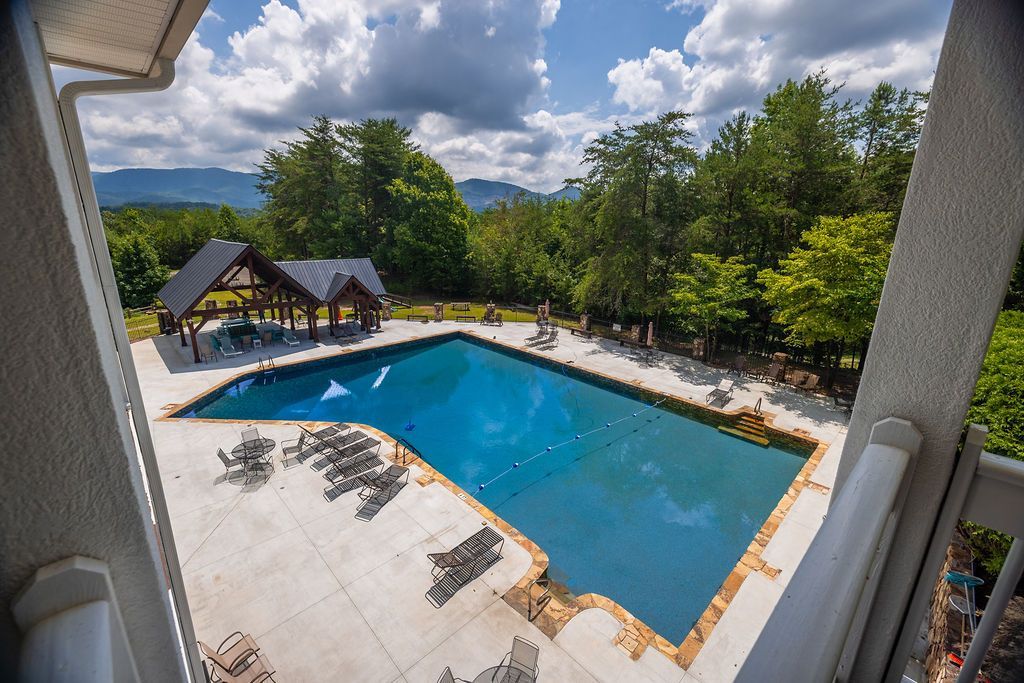 An aerial view of a blue outdoor pool with lounge chairs and a gazebo, surrounded by trees and mountains.