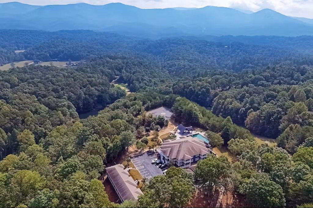Aerial view: Large house with pool in forest, mountains in background.