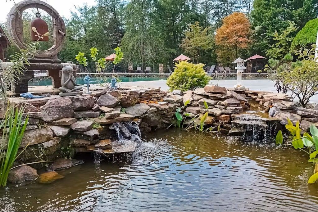 A serene garden scene with a small waterfall cascading into a pond. Trees, a pool and a bell structure are in the background.