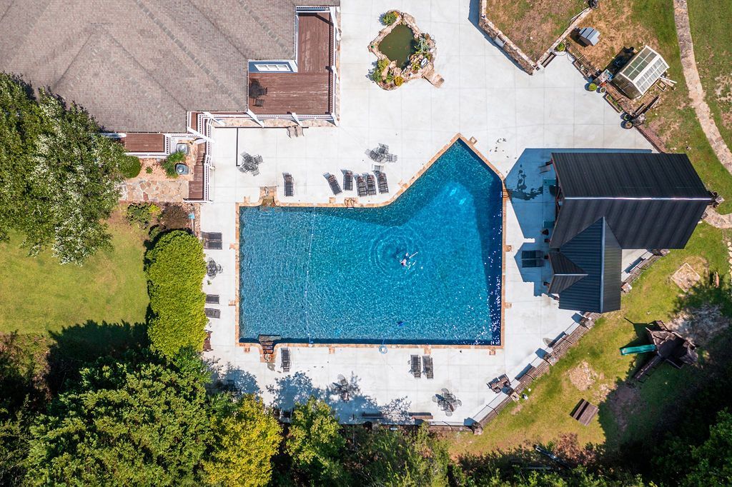 Aerial view: Swimming pool surrounded by concrete, lounge chairs, a building and green areas.