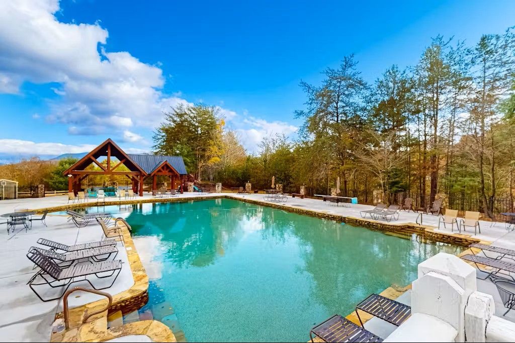 A pool with clear water, surrounded by trees and seating, under a bright blue sky.