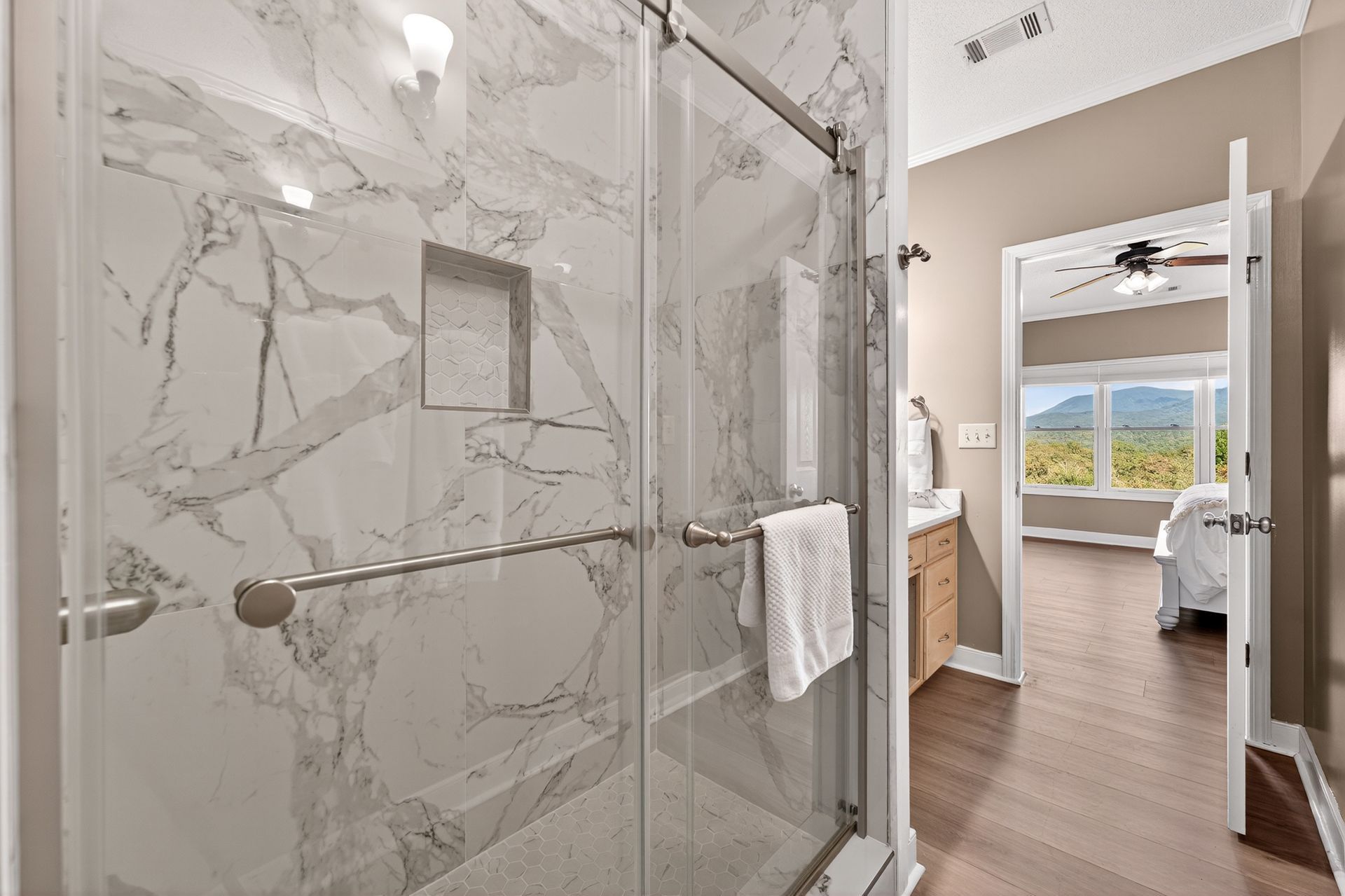 Bathroom with marble-tiled shower, glass door, and towel, open to a hallway and bedroom with a view.