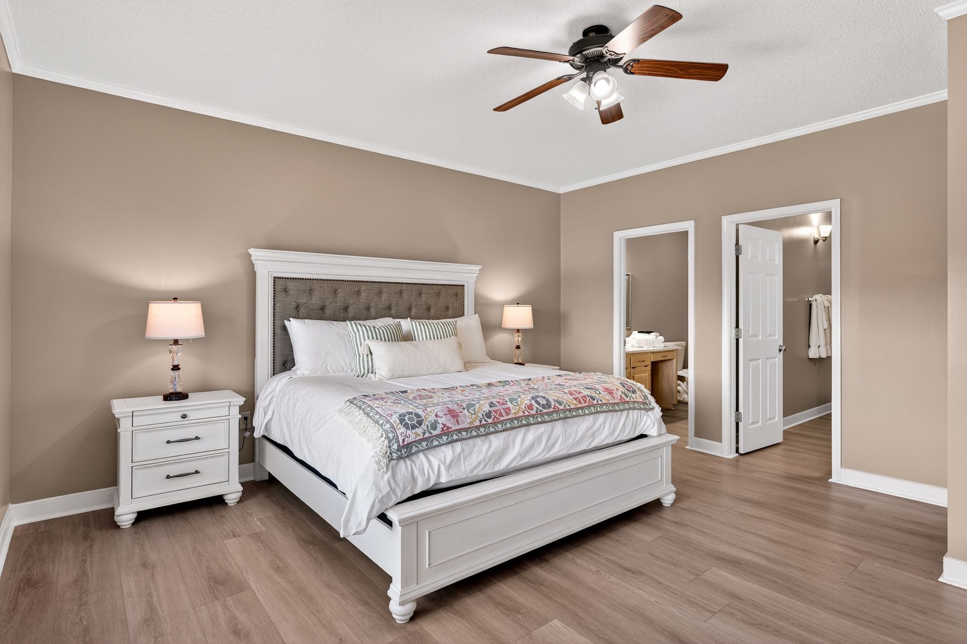 Bedroom with white bed, lamps, and wood-look floors. A door leads to another room.