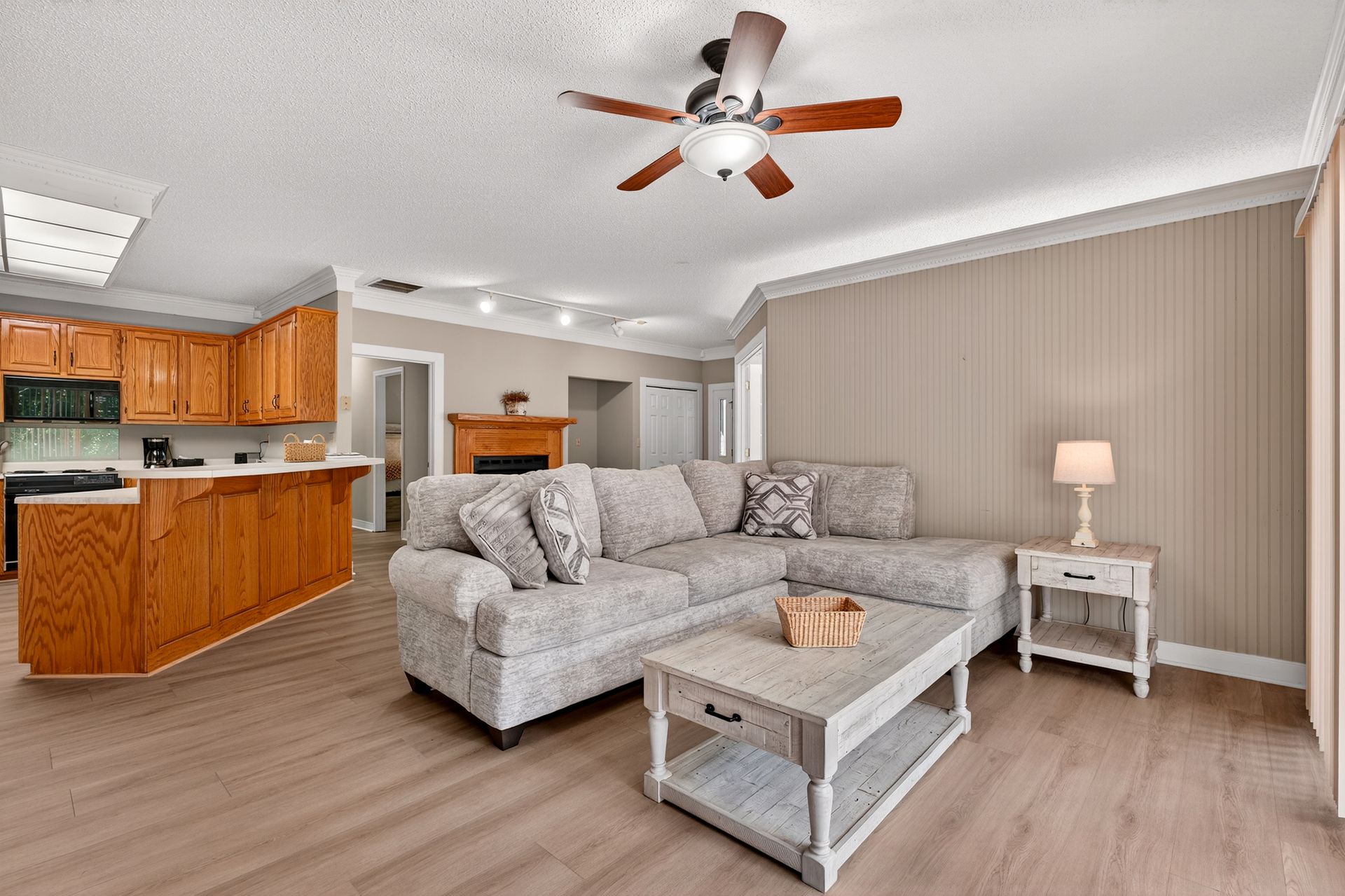 Cozy living room with kitchen, wood cabinets, gray sectional, and light-colored wood floors.
