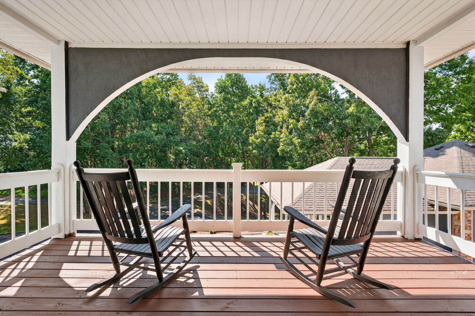 Two black rocking chairs on a wooden porch overlook lush trees through an arched opening.