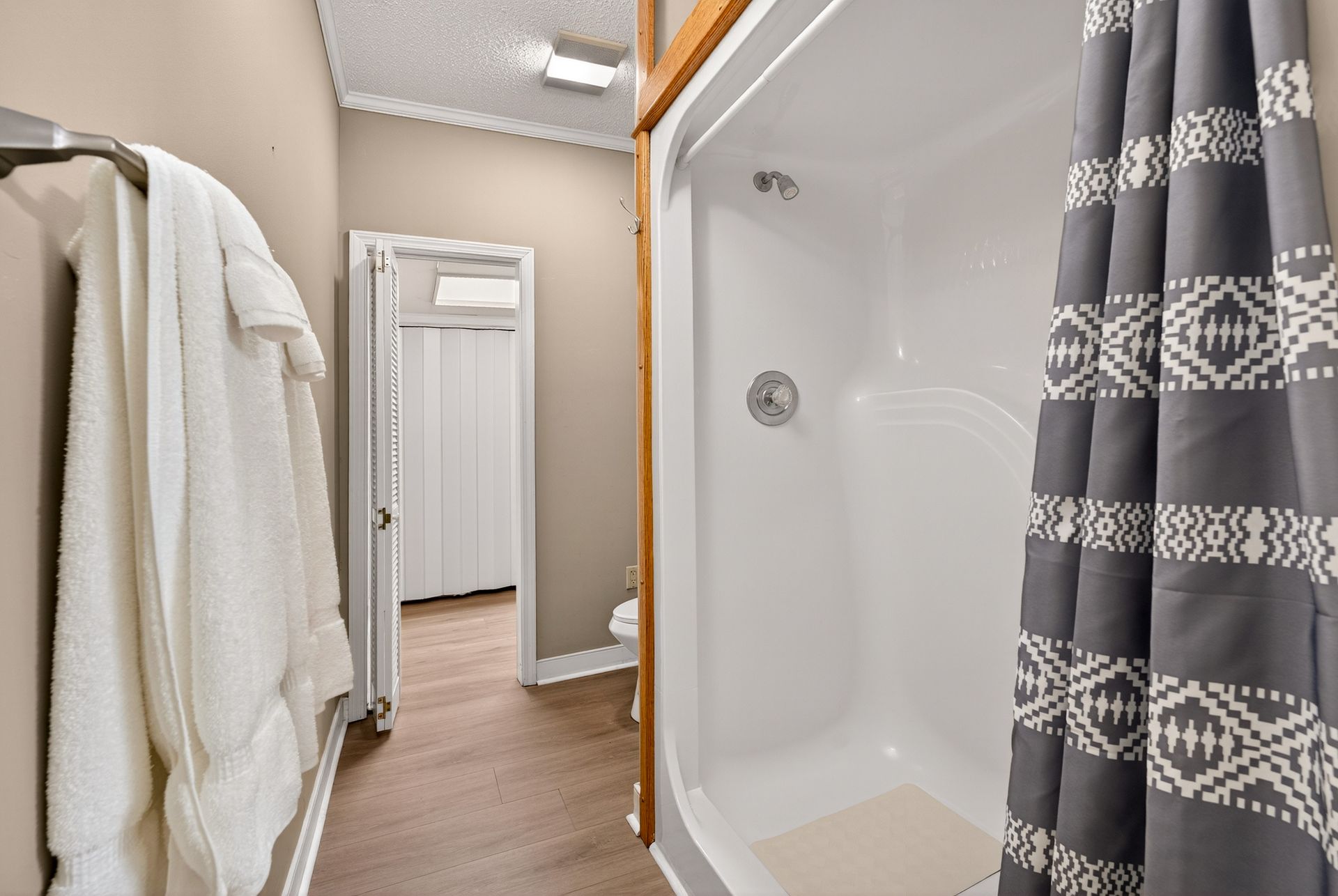 Bathroom with a white shower, patterned gray curtain, and white towels on a rack. A door to another room is open.