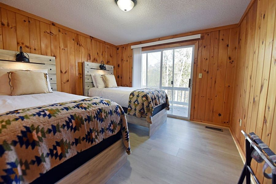 Bedroom with two beds, wood-paneled walls, sliding glass door to a balcony, and light-colored flooring.