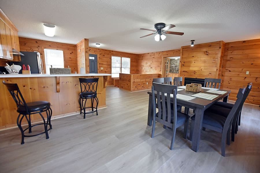 Dining area with wooden walls and gray flooring. Table set for six, two bar stools.