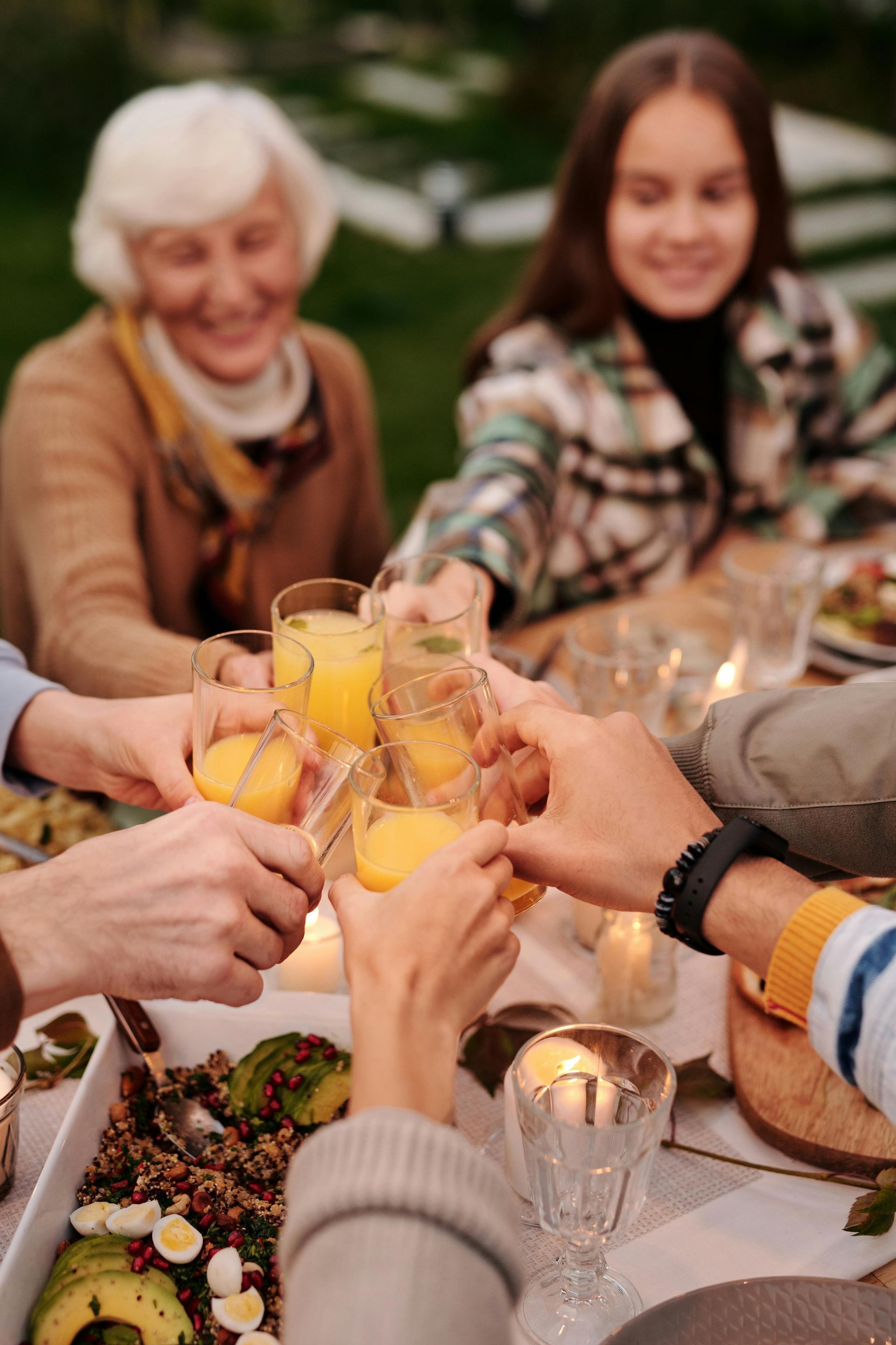Family toasting drinks at an outdoor dinner table, smiling; a senior woman and teenage girl are visible.
