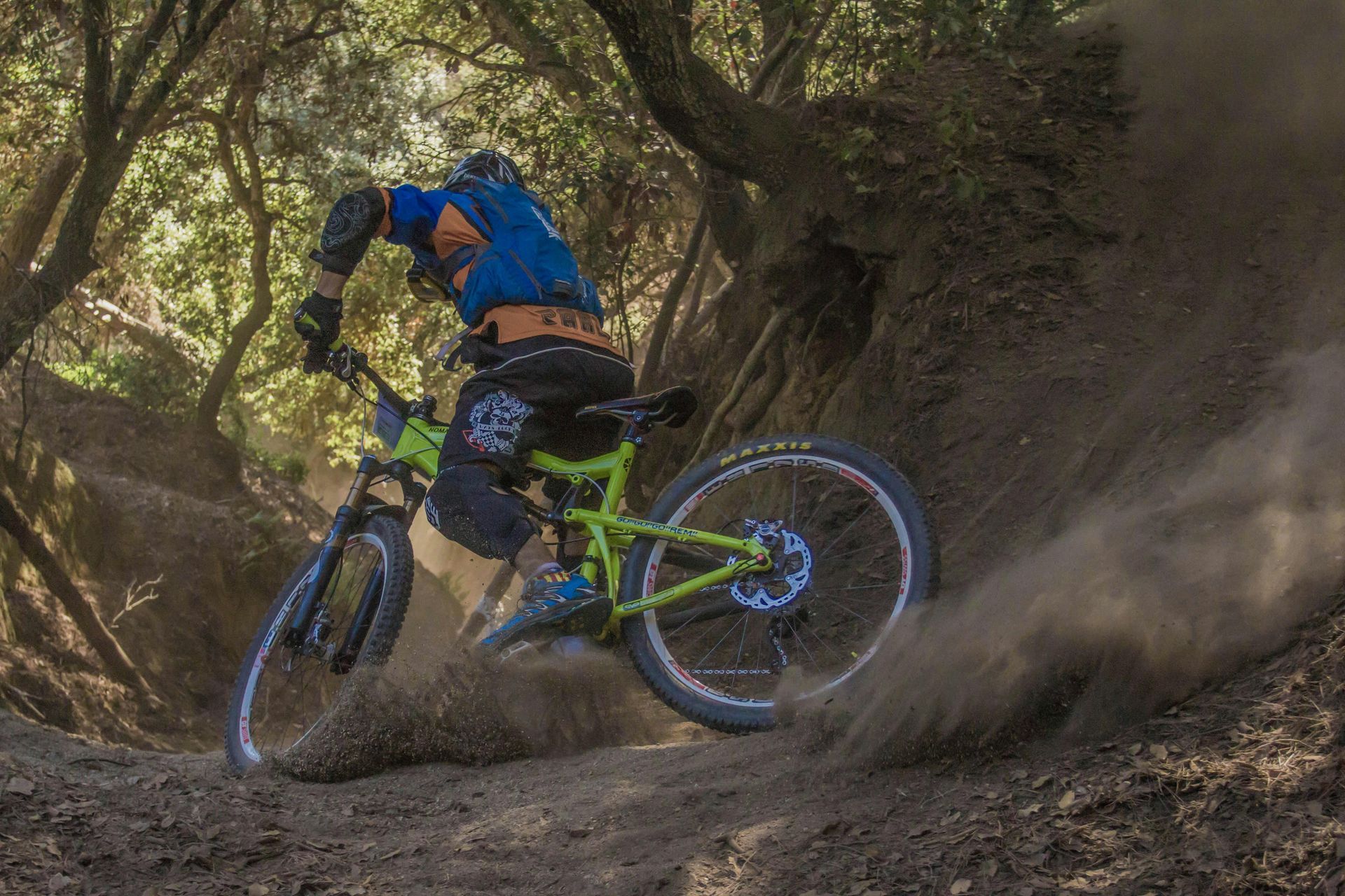Mountain biker in blue and green gear, kicking up dust on a dirt trail in a forest.