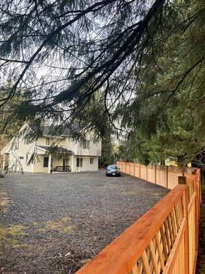 Gravel driveway beside a beige house and blue car, viewed through trees and a wooden fence.