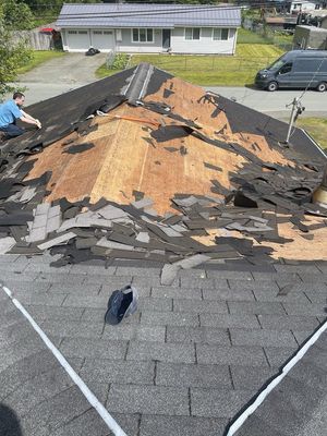 Roof under repair with large exposed plywood, torn shingles, and workers on a residential house