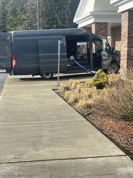 Dark van parked in a driveway beside a brick house, with equipment visible in the open side door.