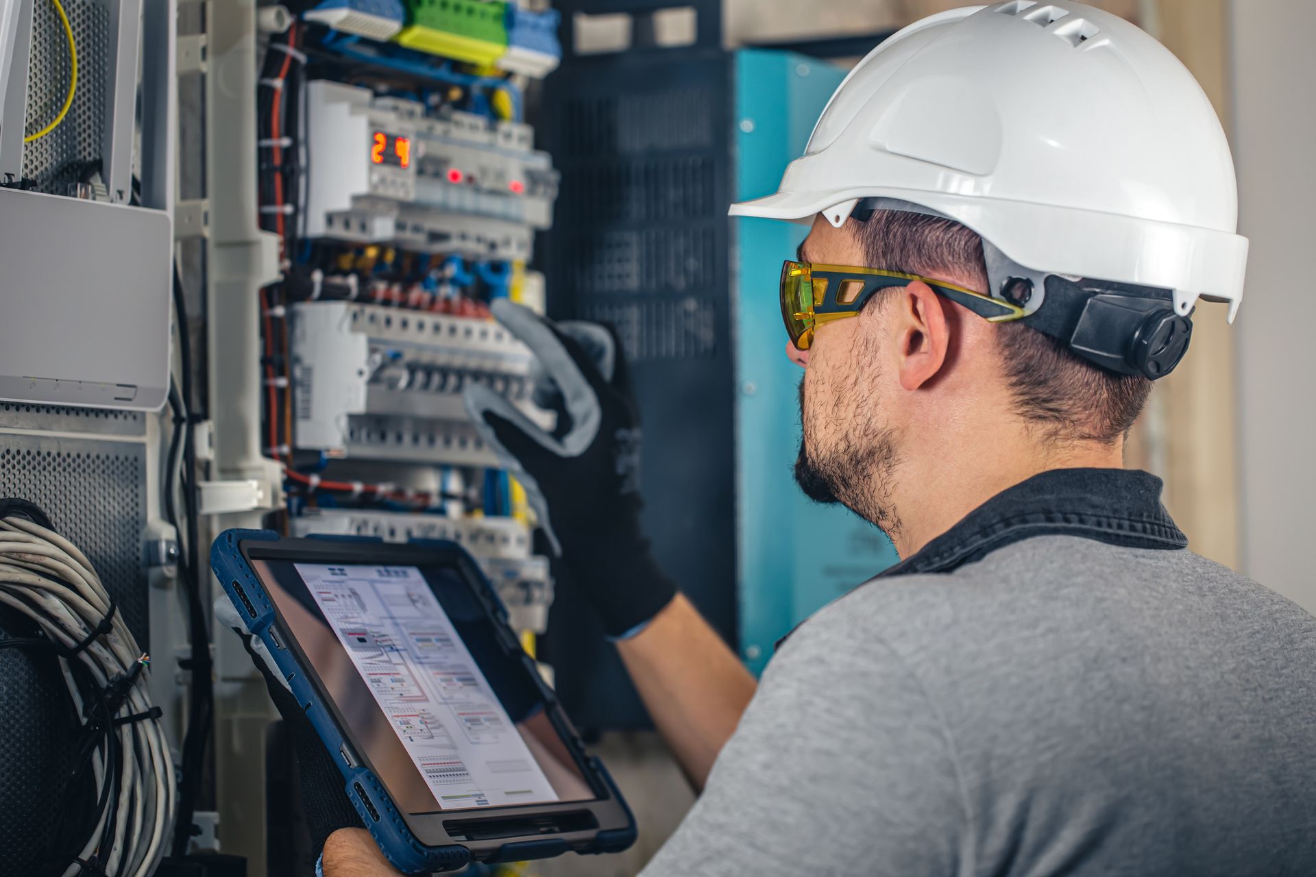 Electrician in hard hat and safety glasses examines electrical panel, using a tablet.