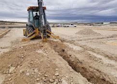 A yellow backhoe digs a trench in a dirt construction site under a cloudy sky.