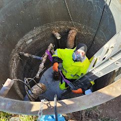 A worker in a high-visibility jacket and respirator mask descends a metal ladder into a circular concrete utility manhole.