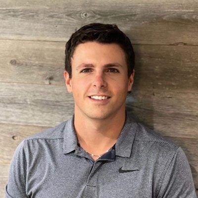 Man with dark hair and a grey polo shirt smiling against a wooden wall.
