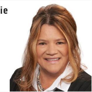 Woman with shoulder-length highlighted hair, smiling, wearing a black blazer and white shirt, against a white background.