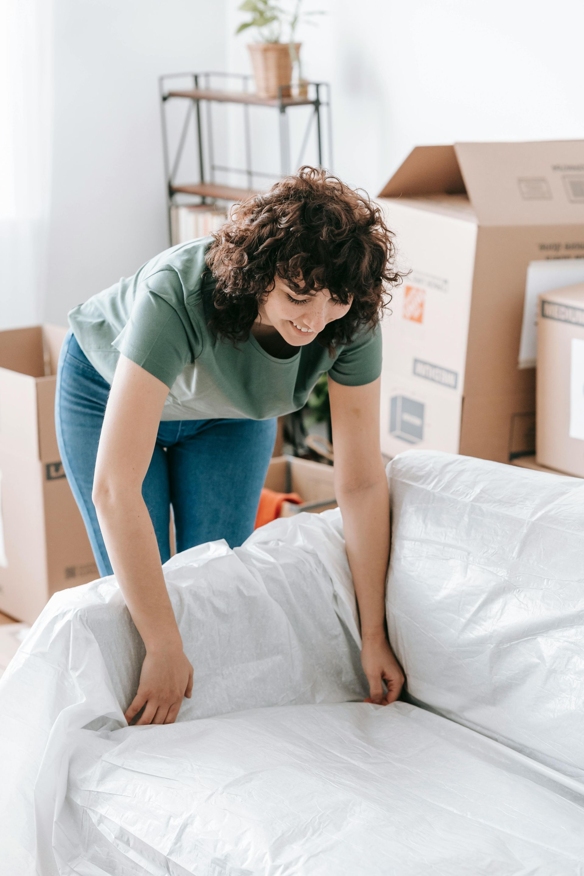 Woman wrapping a couch in plastic, surrounded by moving boxes, in a room.