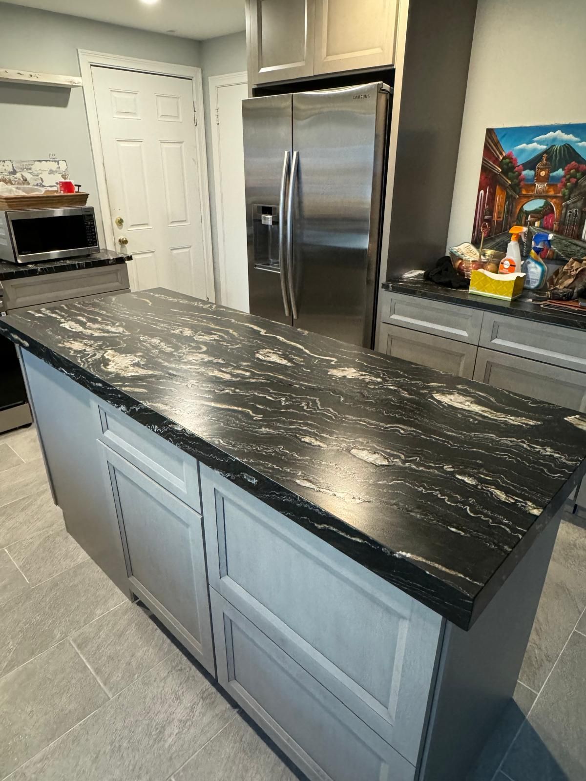 Kitchen island with black and cream speckled countertop; light gray cabinets; stainless steel fridge.