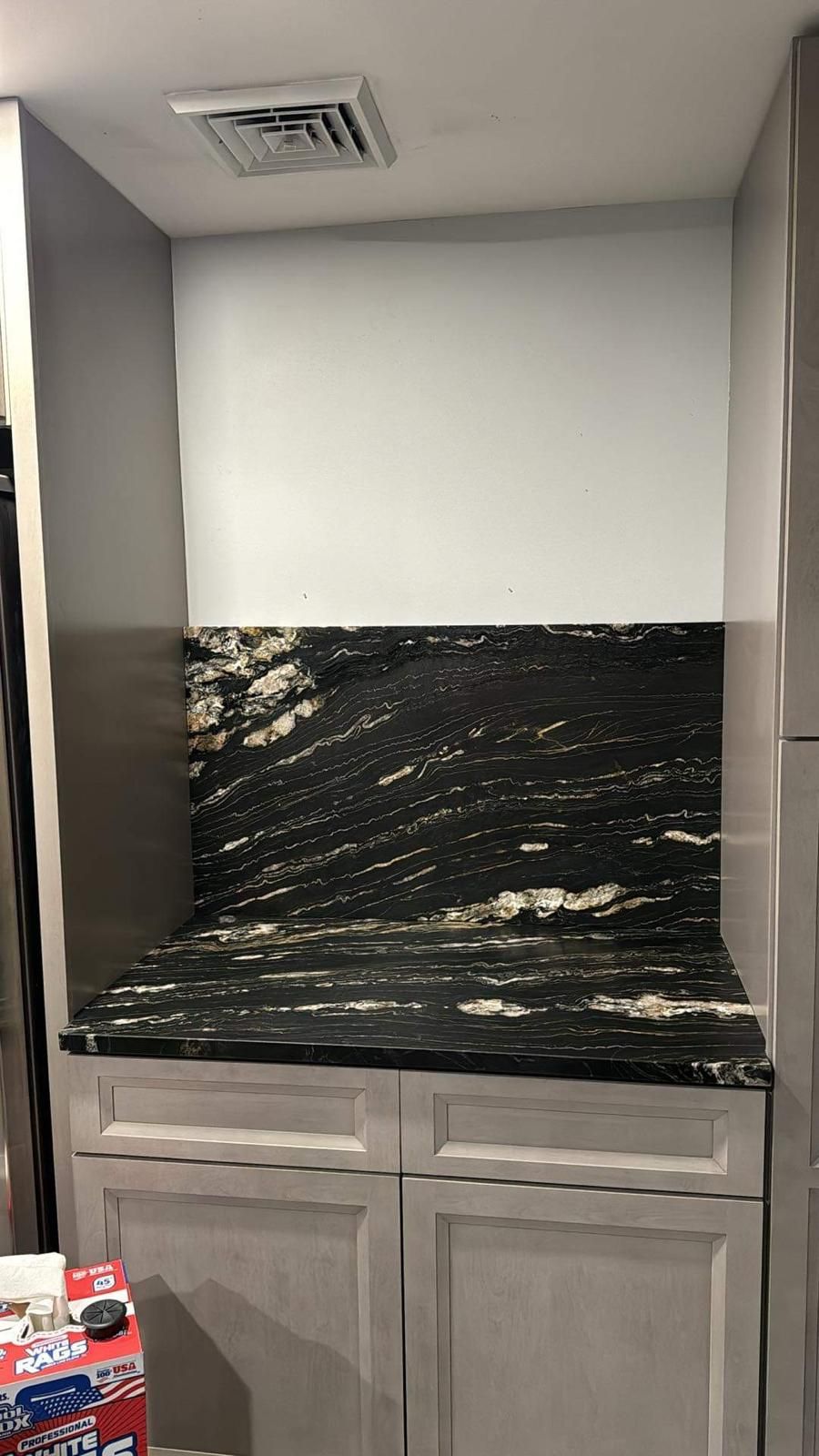 Built-in cabinet with granite countertop and backsplash, above light-colored lower cabinets, gray walls, and vent.