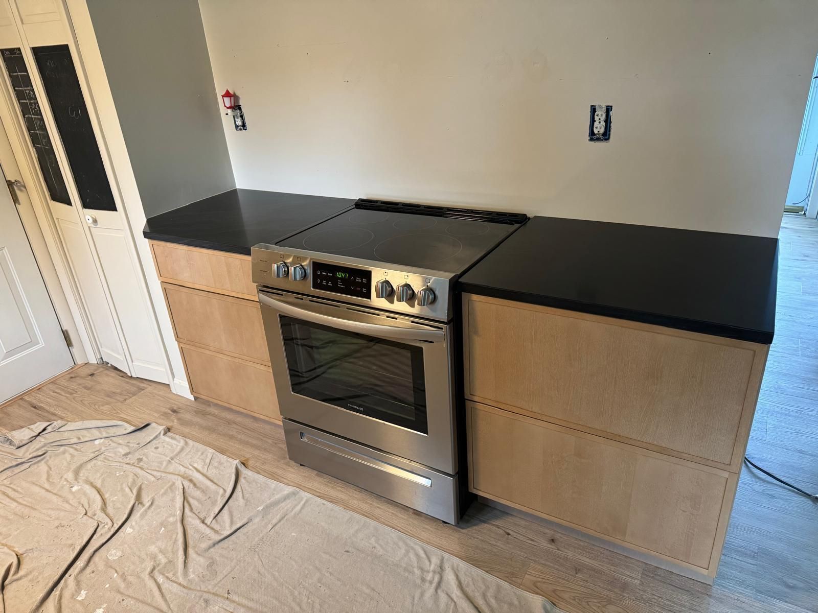 Kitchen with stainless steel range, black countertop, and light wood cabinets against a gray wall.