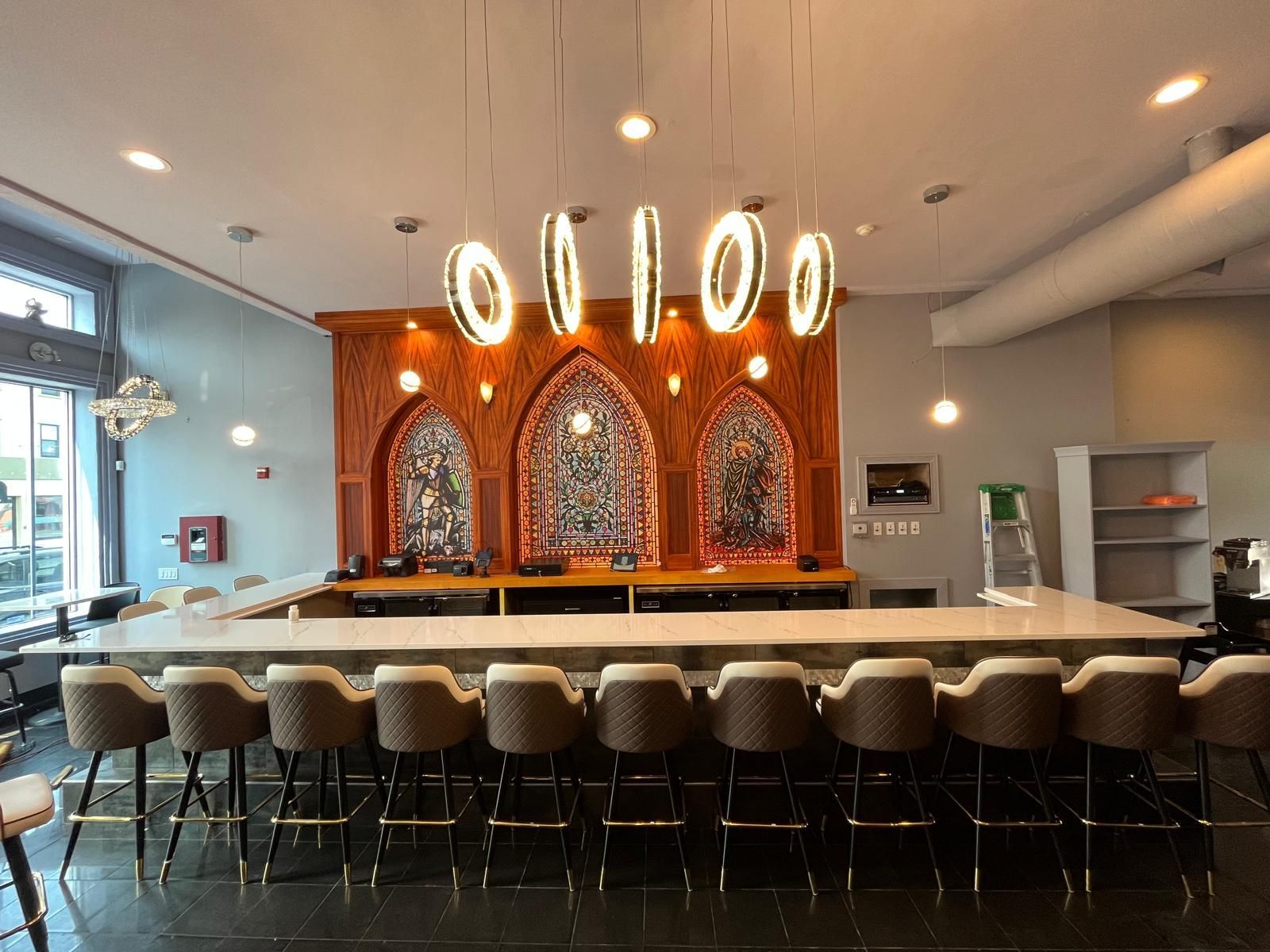 A bar with a long counter and stools. Ornate lighting hangs above. Wooden backdrop with stained glass.