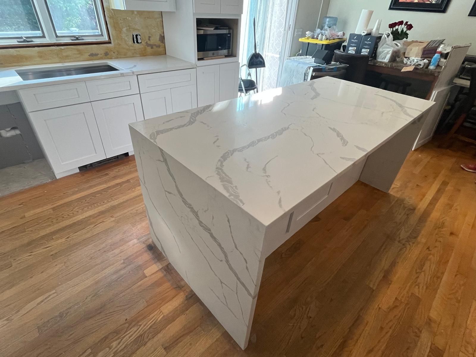 White kitchen island with veined countertop, on wood floor. White cabinets and cooktop in background.