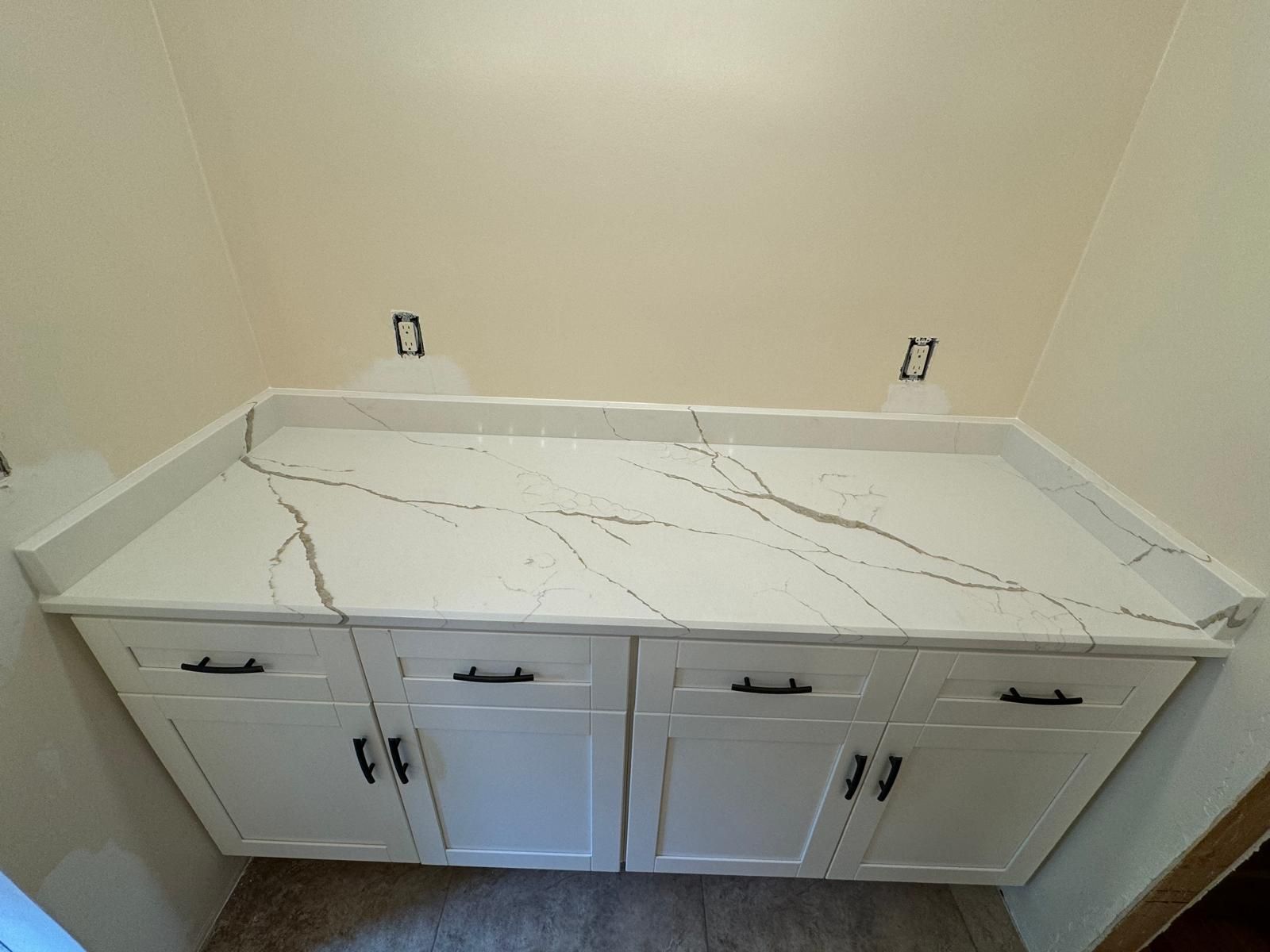 White cabinets with dark handles, topped by a white quartz countertop with gray veining. Beige wall in the background.